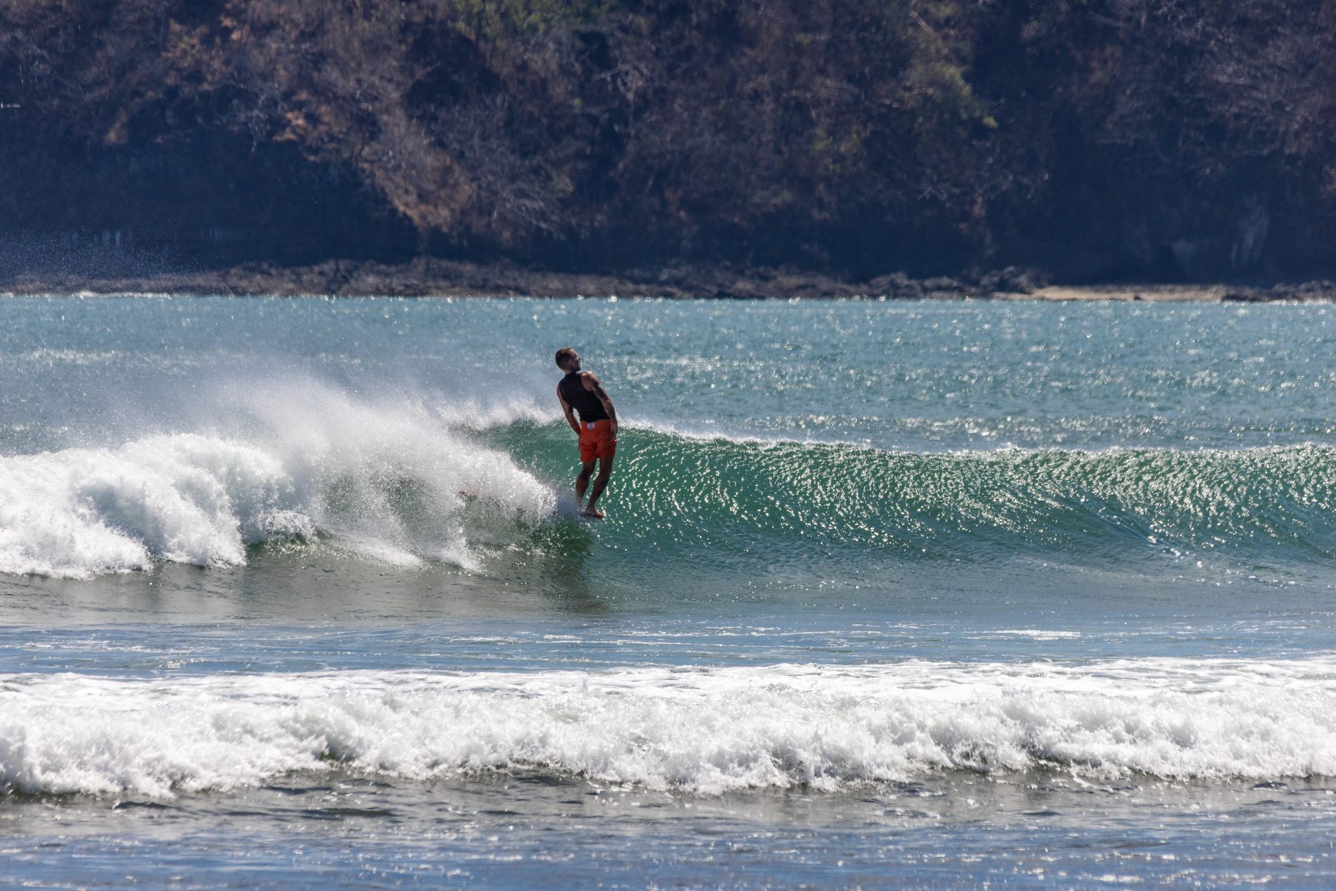 A surfer riding a green wave along a jungle-covered headland